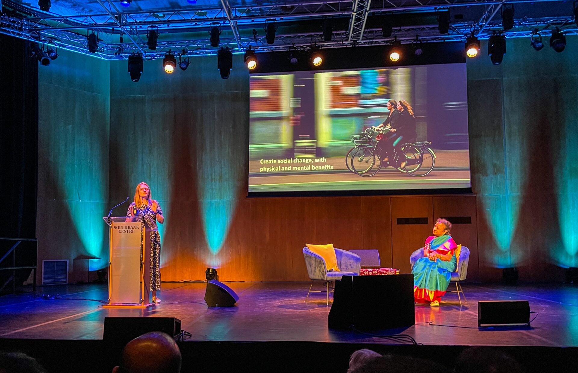 Two people on a stage, one at a lectern talking, the other sat on a chair. Presentation with image of people cycling in the backgroud.